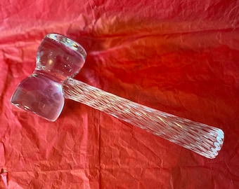 May include: A clear glass hammer with a textured handle and a hammerhead shape. The hammer is lying on a red, textured surface.
