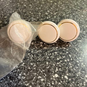 May include: Three white cabinet knobs with gold trim. One knob is still in its clear plastic packaging. The knobs are round and have a simple, elegant design. They are likely used for drawers or cabinets.