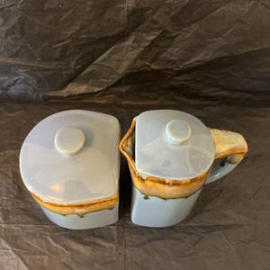 May include: A light blue ceramic creamer and sugar bowl set. The set has a rounded design with a lid and a handle on the creamer. The bottom of the set has a brown and tan color.