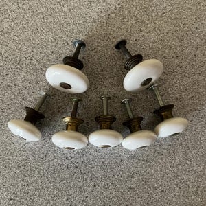 May include: A set of six white ceramic cabinet knobs with round, smooth tops and metallic, brass-toned bases. Each knob features a central circular metal inlay on the top and includes a threaded metal screw extending from the back for installation.