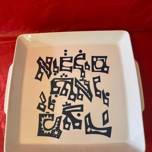 May include: A white square serving tray with handles, featuring a dark blue abstract design in the center. The tray is set against a red background. The text on the tray is stylized and artistic.