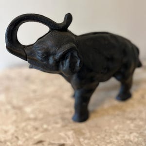 May include: A black cast iron elephant figurine with a raised trunk and a slot on its back. The elephant has a textured surface and is standing on a light-colored surface.