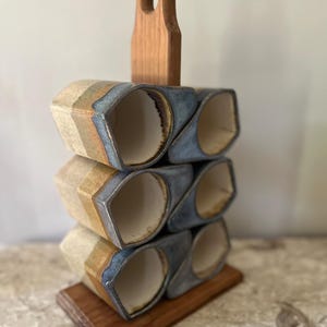 May include: A handmade ceramic wine rack with a wooden handle and base. The rack holds six hexagonal wine glass holders. The holders are a mix of beige, blue, and brown. The wooden handle has a hole at the top for easy carrying.