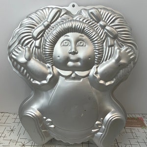 Può includere: Uno stampo per torta in metallo argentato a forma di bambola Cabbage Patch Kid. La bambola ha un viso rotondo con guance rosate, occhi blu e un fiocco rosso nei capelli.
