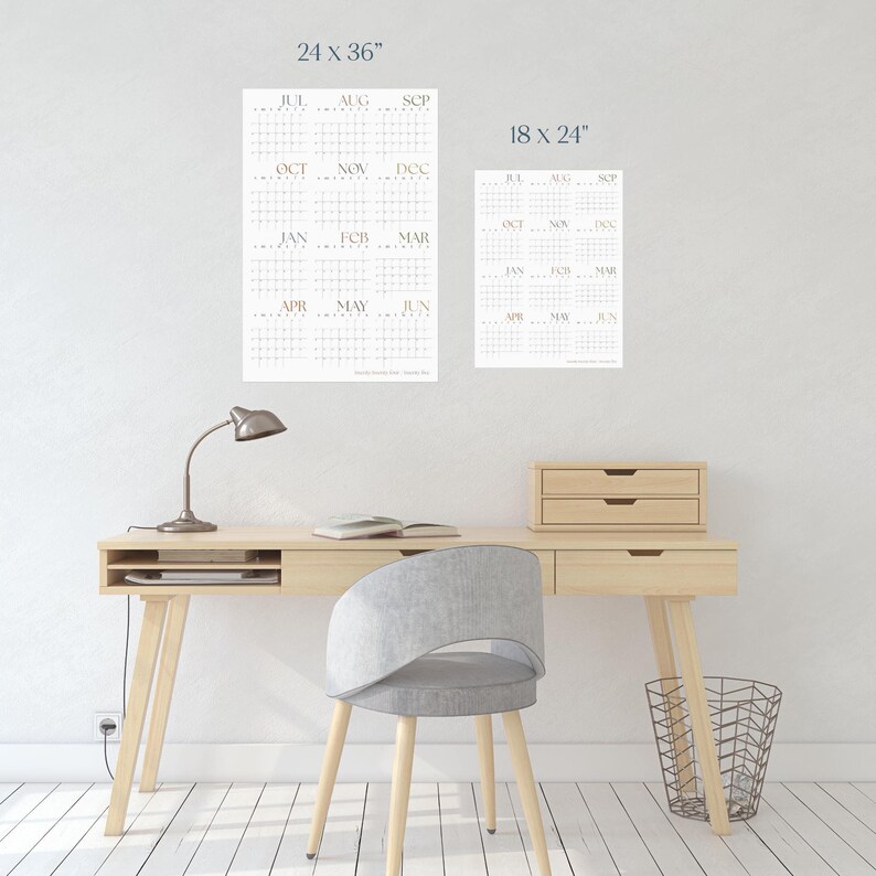 May include: Two white calendar posters with a grid layout and months of the year printed in gold. The larger poster is 24 inches by 36 inches and the smaller poster is 18 inches by 24 inches. The posters are hanging on a white wall above a wooden desk with a lamp, chair, and wastebasket.