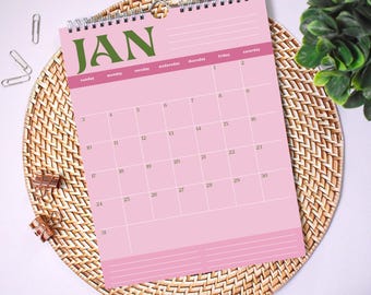 May include: A wall calendar for the month of January featuring a light pink background with dark green text reading JAN. The grid displays days of the week and dates from 1 to 31 in dark green, with white grid lines and lined sections for notes.