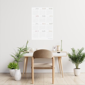 May include: A white wall with a calendar hanging on it. The calendar is a grid with each month labeled in a light pink font. The calendar is hanging above a wooden desk with a brown chair. There are two potted plants on the floor.
