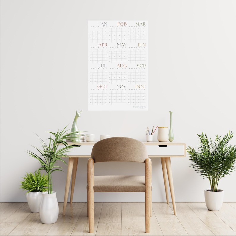May include: A white wall with a calendar hanging on it. The calendar is a grid with each month labeled in a light pink font. The calendar is hanging above a wooden desk with a brown chair. There are two potted plants on the floor.