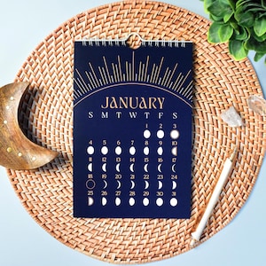 May include: A navy blue wall calendar for January, featuring a gold sunburst design and moon phase illustrations. The calendar is spiral-bound and rests on a woven placemat. A white pen and decorative items are also present.