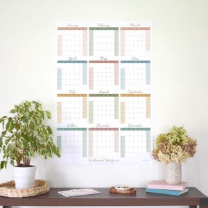 Op de afbeelding: Een wandkalender met een minimalistisch ontwerp met een raster van 12 maanden, elk met een andere pastelkleur. De maanden zijn in cursief schrift gelabeld. De kalender heeft de titel "Twee Duizend Eenentwintig".
