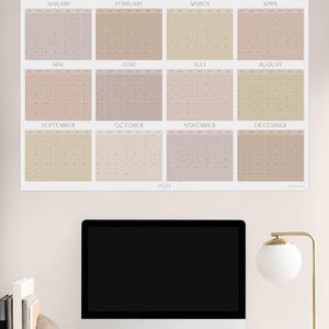 May include: A beige and light brown calendar for the year 2023, with each month displayed in a separate square. The calendar is hanging on a light beige wall above a computer monitor and a desk lamp.