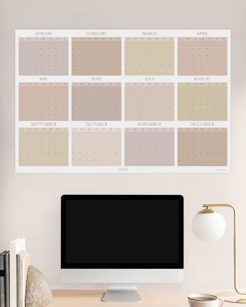 May include: A beige and light brown calendar for the year 2023, with each month displayed in a separate square. The calendar is hanging on a light beige wall above a computer monitor and a desk lamp.