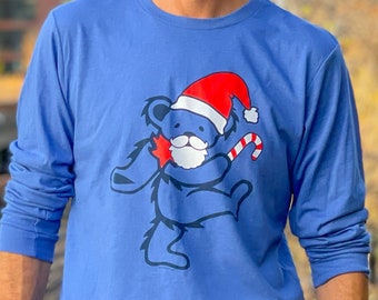 May include: A long-sleeved blue shirt featuring a dancing bear design. The bear is wearing a Santa hat and holding a candy cane. The bear has a white beard and a red star. The shirt is made of a soft material.
