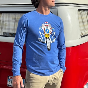 May include: A blue long-sleeve t-shirt with a graphic of a skeleton riding a motorcycle. The skeleton is wearing a red, white, and blue hat and holding a flag. The motorcycle has a yellow wheel and a blue and white stripe.