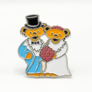Könnte beinhalten: Emaille-Anstecknadel mit zwei lächelnden Bären in Hochzeitskleidung. Einer der Bären trägt einen Zylinder und einen blauen Anzug, der andere ein weißes Kleid und hält einen roten Blumenstrauß. Die Bären haben gelbes Fell und schwarze Akzente.
