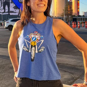 May include: A light blue tank top with a graphic of a cartoon skeleton riding a motorcycle. The skeleton is wearing a red, white, and blue hat and sunglasses. The tank top is cropped and has a racerback style.