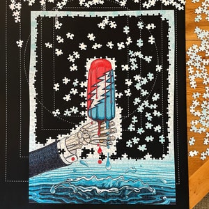 May include: A completed jigsaw puzzle featuring a red, white, and blue popsicle held by a skeleton hand. The popsicle has a lightning bolt design. The puzzle is set against a black background with scattered puzzle pieces.