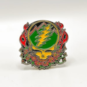 May include: A colorful enamel pin featuring a yellow skull with a lightning bolt and mountain design. The pin has a green and yellow lightning bolt, surrounded by a green mountain range, red flames, and a border of red roses.