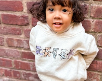 Puede incluir: Un niño pequeño con una sudadera con capucha de color crema con un gráfico de cinco osos bailando de colores. La sudadera tiene una capucha con cordón y mangas largas. El niño lleva pantalones vaqueros azules claros.