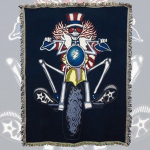 May include: A woven tapestry featuring a skeleton wearing a red, white, and blue Uncle Sam hat riding a motorcycle. The motorcycle has a large front tire and the skeleton is wearing sunglasses.