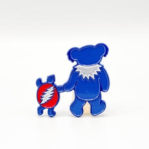 May include: A blue bear-shaped pin with a silver glitter collar and a smaller red turtle-shaped pin. The turtle pin features a white lightning bolt design. Both pins are on a white background.