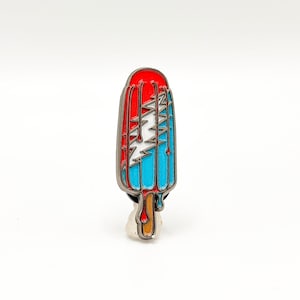 May include: A colorful enamel pin shaped like a popsicle. The pin features a red top, blue stripes, and a white lightning bolt design. The popsicle stick is brown, and the pin has a silver border.