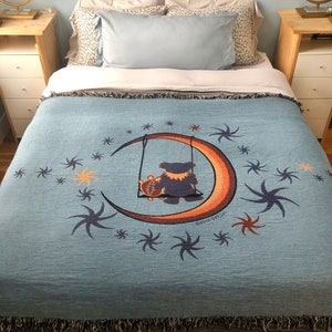 May include: A blue woven throw blanket with a crescent moon and two bears on a swing. The moon is orange and yellow and the bears are dark blue. The background is a light blue with white stars.