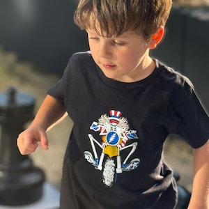 May include: Black t-shirt featuring a colorful graphic of a motorcycle with a skull wearing a hat. The design includes red, white, blue, and yellow elements. The shirt is worn by a young child.