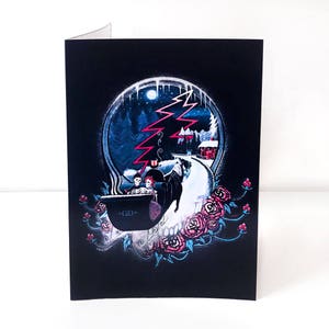 May include: A black greeting card featuring a skull design with a winter scene inside. The scene includes a road, trees, and a red building. Pink and blue roses surround the skull. The card has the initials "GD" on the bottom left.