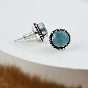 Puede incluir: Pendientes de botón con una piedra preciosa redonda de color azul claro engastada en metal plateado. Los pendientes tienen un diseño de cuerda trenzada alrededor de la piedra preciosa y un cierre de poste. Se muestran sobre una superficie blanca.
