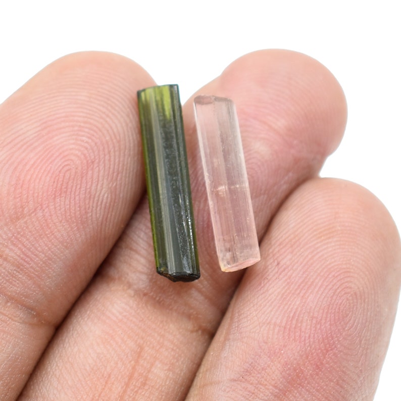 May include: Two tourmaline crystals, one olive green and the other a pale pink, are displayed. The crystals are rectangular and have a translucent appearance. The background is a plain white, highlighting the natural beauty of the gemstones.