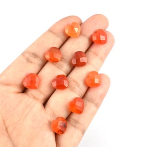 May include: Eight small, faceted heart-shaped carnelian gemstones in shades of orange and red. Each stone features a polished, multi-faceted surface that reflects light, showcasing the natural translucent quality and warm color variations of the mineral.