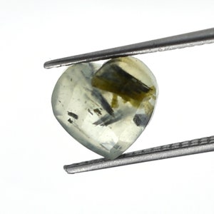 May include: A heart-shaped, translucent pale yellow gemstone featuring visible dark green and black needle-like inclusions throughout its structure. The stone is held by a pair of metal tweezers, showcasing its faceted surface and internal mineral patterns.