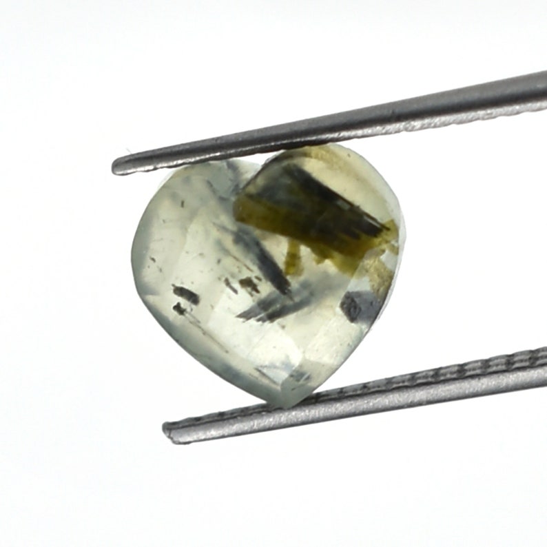 May include: A heart-shaped, translucent pale yellow gemstone featuring visible dark green and black needle-like inclusions throughout its structure. The stone is held by a pair of metal tweezers, showcasing its faceted surface and internal mineral patterns.