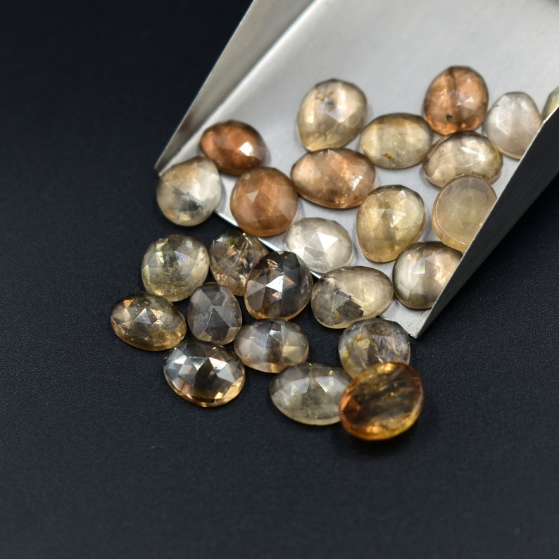 May include: A collection of oval-shaped, faceted gemstones in shades of brown, gray, and clear. The stones are arranged in a pile on a silver scoop.