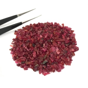 May include: A collection of small, raw, deep red ruby gemstone fragments arranged in a circular pile. The stones exhibit irregular, jagged shapes and varying shades of crimson and maroon. A pair of black metal tweezers is positioned nearby.