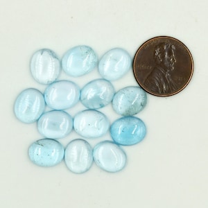 Op de afbeelding: Een verzameling ovaalvormige, lichtblauwe edelstenen. De edelstenen variëren in tint, sommige lijken bijna wit en andere dieper blauw. Een munt is inbegrepen ter vergelijking. De edelstenen liggen op een witte ondergrond.