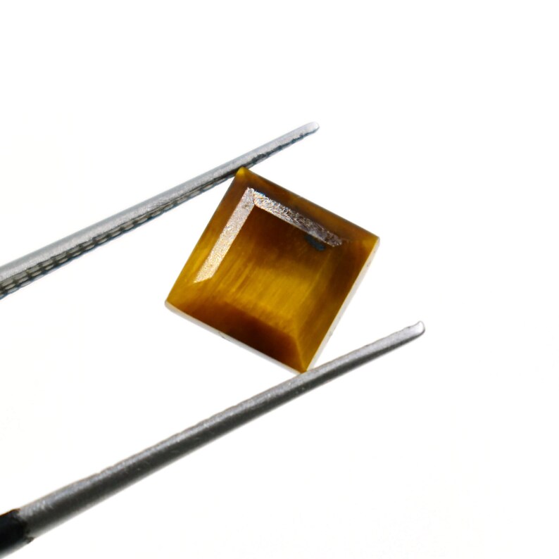 May include: A square-cut, golden-brown tiger's eye gemstone is held by silver tweezers against a white backdrop. The gemstone has a polished surface with visible chatoyancy, reflecting light in a way that resembles a cat's eye.