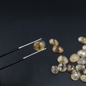 May include: A collection of oval-shaped, faceted, light brown gemstones. One gemstone is held in tweezers, while the others are scattered on a black surface.