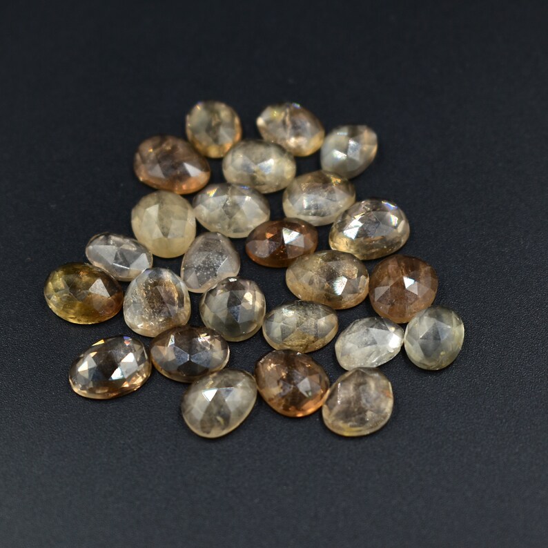 May include: A collection of 20 faceted oval-shaped gemstones in shades of brown and clear. The stones are arranged in a loose pile on a black background.