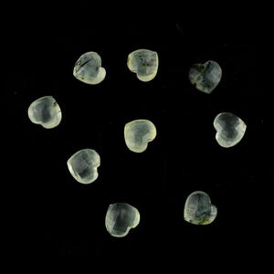May include: Nine small, faceted heart-shaped gemstones with a translucent, pale green hue and subtle internal inclusions. Each stone features a polished surface with multiple facets that catch the light, arranged in a scattered pattern.