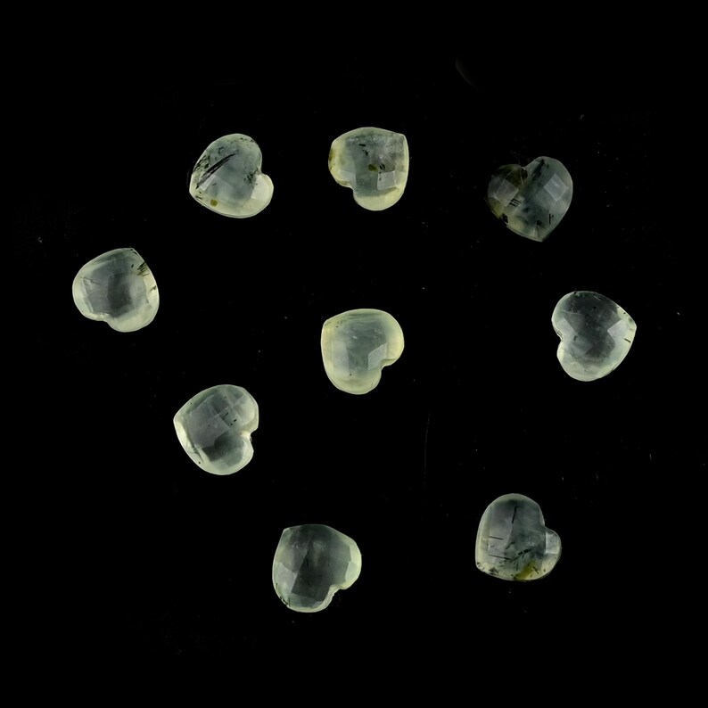 May include: Nine small, faceted heart-shaped gemstones with a translucent, pale green hue and subtle internal inclusions. Each stone features a polished surface with multiple facets that catch the light, arranged in a scattered pattern.