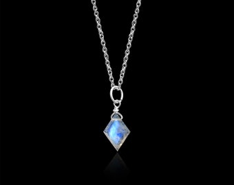 Puede incluir: Un collar de cadena plateada con un colgante en forma de diamante que contiene una piedra lunar azul translúcida e iridiscente. La piedra está asegurada con un alambre plateado en la parte superior, unido a una argolla circular en la cadena.