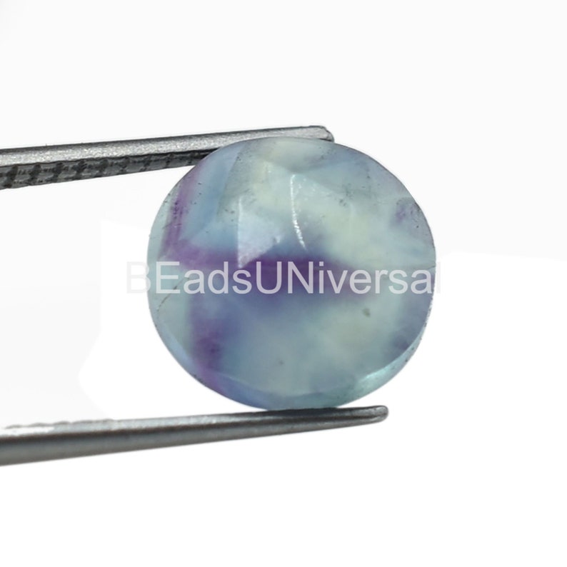 May include: A round, light blue fluorite gemstone with purple and white streaks. The gemstone is held by a pair of tweezers.