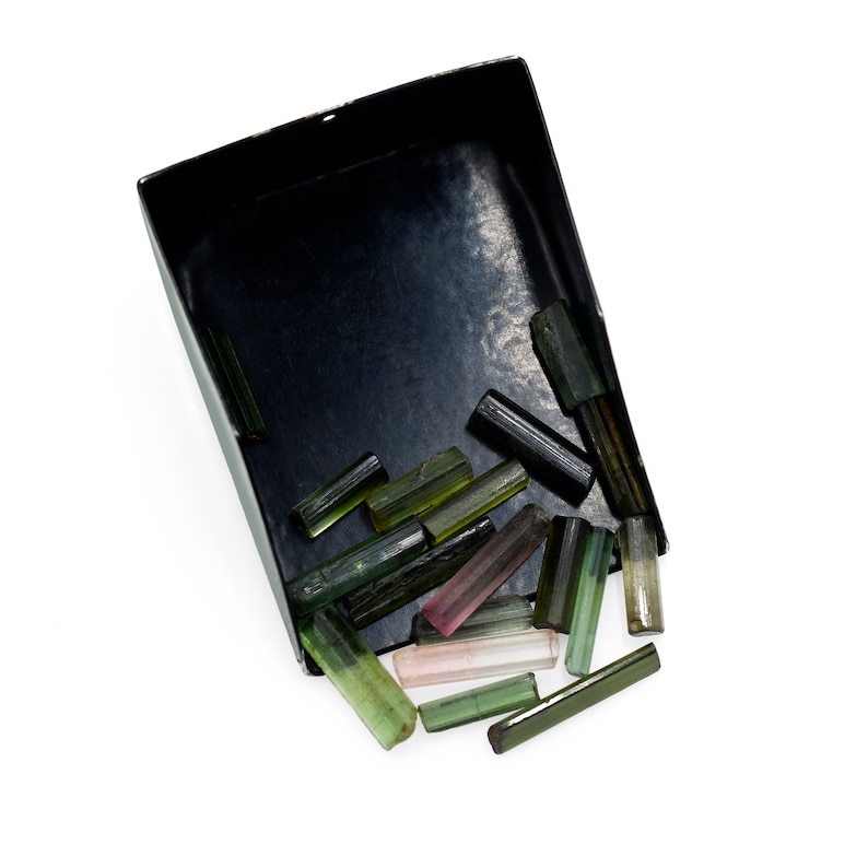 May include: A black rectangular tray filled with raw tourmaline crystals. The crystals vary in color, including green, pink, and black. The crystals are in various sizes and shapes, with a rough, natural texture.