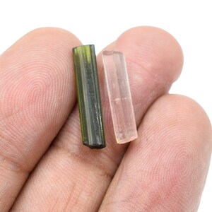 May include: Two tourmaline crystals, one olive green and the other a pale pink, are displayed. The crystals are rectangular and have a polished appearance. The background is a plain white, highlighting the natural beauty of the gemstones.