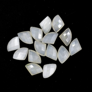 Puede incluir: Una colección de 14 cabujones de piedra preciosa facetados, iridiscentes y blancos. Los cabujones tienen forma de media luna y están dispuestos en un patrón aleatorio.
