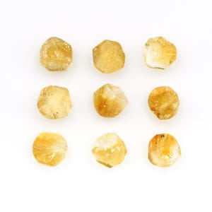 May include: Nine rough-cut, golden-yellow citrine gemstones are arranged on a white background. The stones vary slightly in shape and size, with a natural, unpolished appearance. The color ranges from pale yellow to a deeper amber hue, with some stones showing internal crystalline structures.