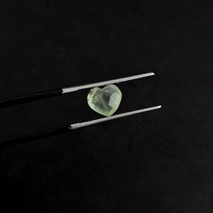 May include: A heart-shaped, pale yellow gemstone with visible dark needle-like inclusions held by a pair of black metal tweezers. The faceted stone displays a translucent quality and a distinct heart silhouette, showcasing its natural internal patterns.