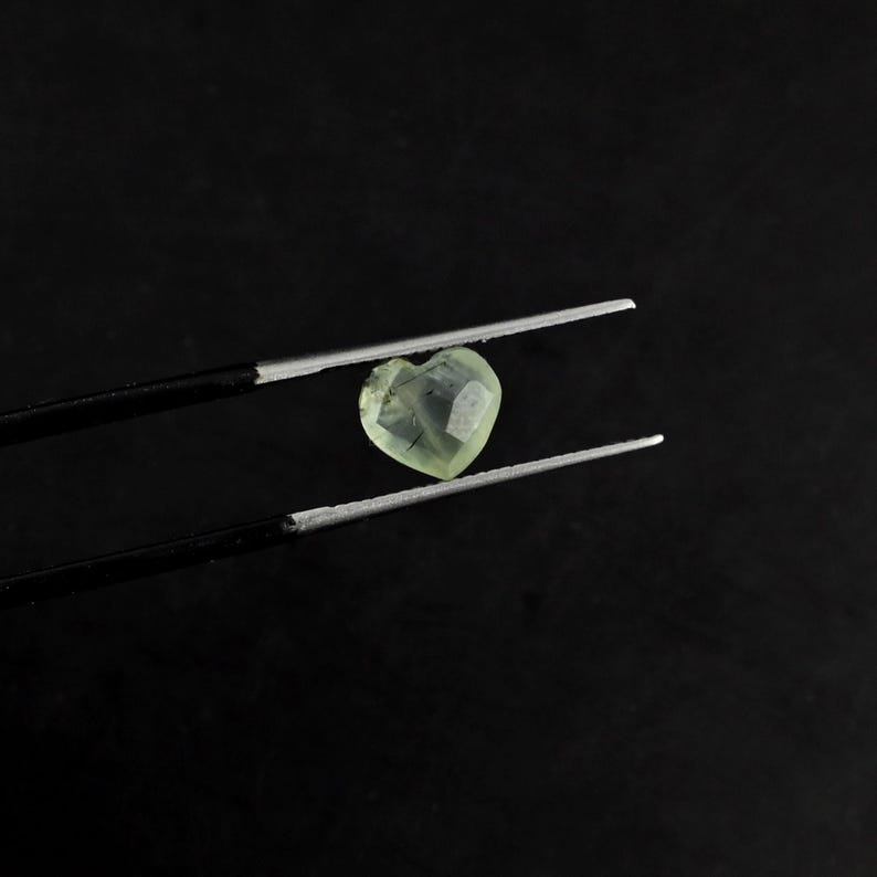 May include: A heart-shaped, pale yellow gemstone with visible dark needle-like inclusions held by a pair of black metal tweezers. The faceted stone displays a translucent quality and a distinct heart silhouette, showcasing its natural internal patterns.